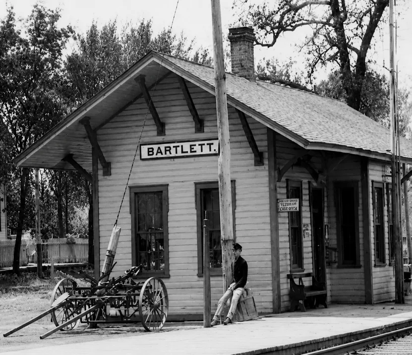 Historic photograph of the wooden Bartlett railroad station, with a man seated on the platform beside a farm cart