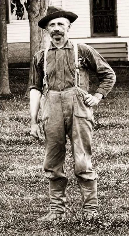 Sepia photograph of J. P. Mason, a moustached farmer in overalls, suspenders and tall boots, standing on the grass outside a farmhouse