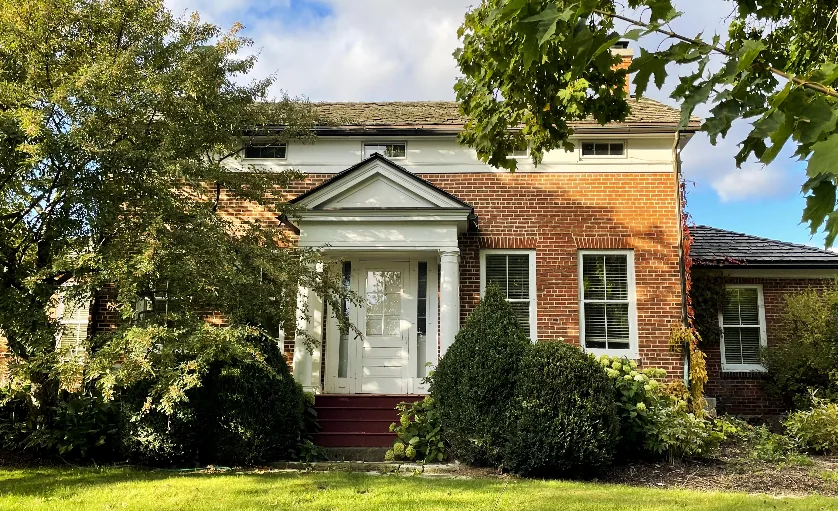 Two-storey red-brick house with a white columned front porch, surrounded by trees and shrubs