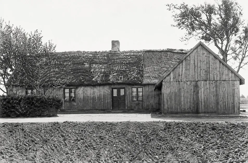 Black-and-white photograph of a long, low thatched farmhouse with a wooden barn at one end, behind a ploughed field in Allerum, Sweden
