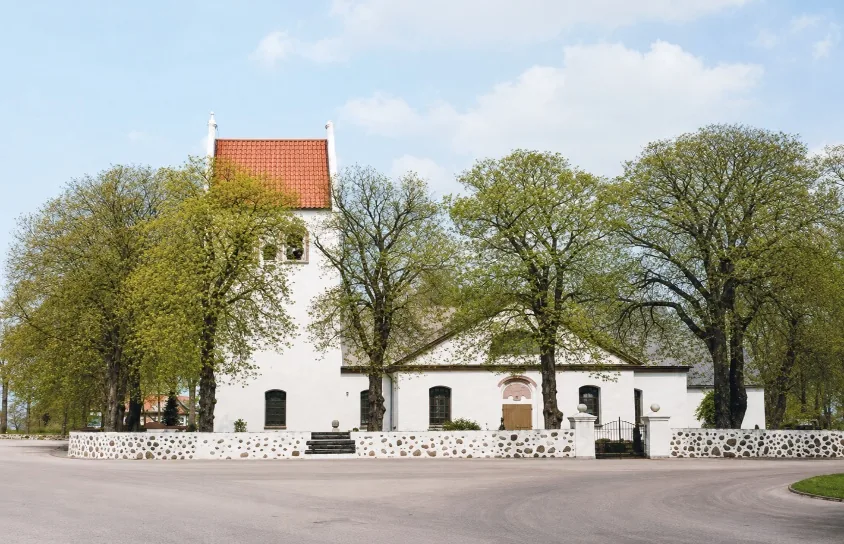 White church with a red-tiled tower surrounded by leafy trees and a low stone wall